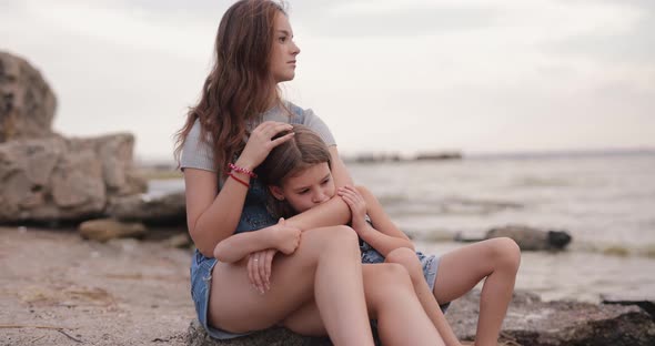 Two Cute Sisters Sitting at the Beach of the Sea and Relaxing