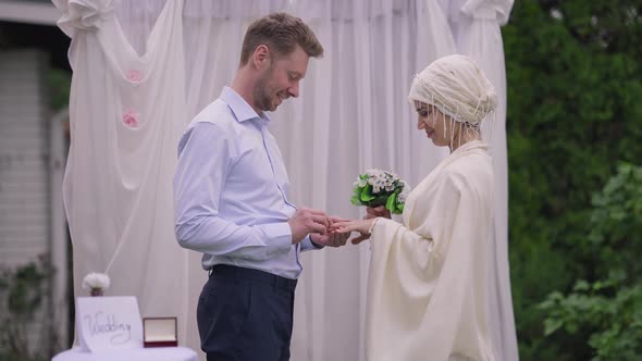 Side View of Happy Middle Eastern Bride Looking with Love at Caucasian Groom Putting Ring on Finger