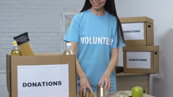 Kind Volunteer Packing Food Supplies in Boxes and Smiling to Camera, Donations