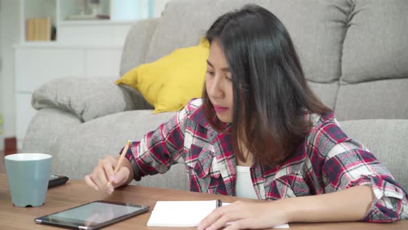 Asian woman do homework at home, female using tablet for searching on sofa in living room at home.
