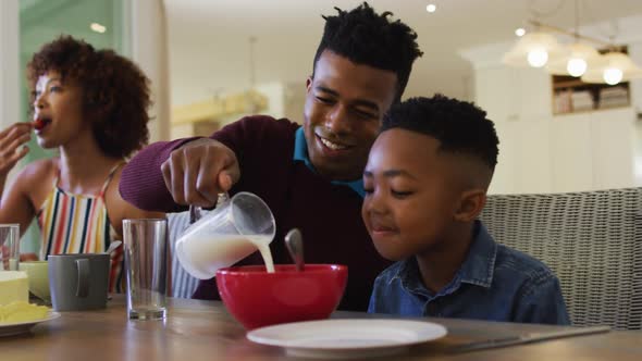 African american father smiling while pouring milk in cereal bowl on his son at home