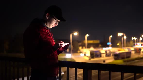 A Young Man Stands on a Bridge Over a Highway and Uses His Smartphone