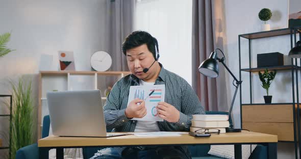 Asian Man in Headset Sitting in Front of Laptop and Explaining Charts to His Partners 