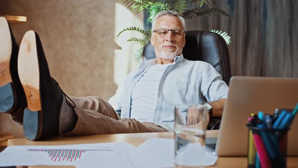 Middleaged Male is Putting Legs on Table Next to Laptop and Smiling Leaning Back in Armchair Working