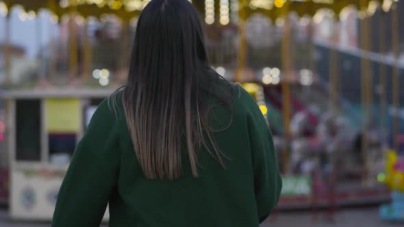 Camera Following Pretty Young Caucasian Woman Walking To Children's Carousels. Cute Girl Turning To