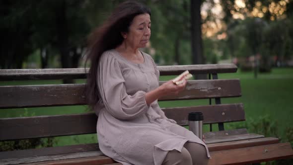 Portrait of Frustrated Senior Caucasian Woman Eating Sandwich Sitting on Bench in Park on Windy