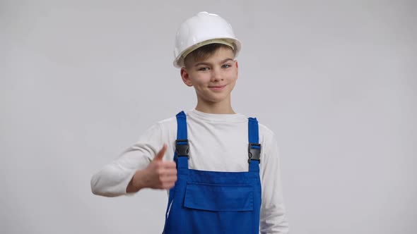 Joyful Caucasian Boy Showing Thumb Up Posing at White Background in Hard Hat and Blue Overalls