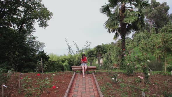 Girl with Smartphone Sitting on Bench on Top of Hill in Tropical Park, Palm Trees, Flower Plantation