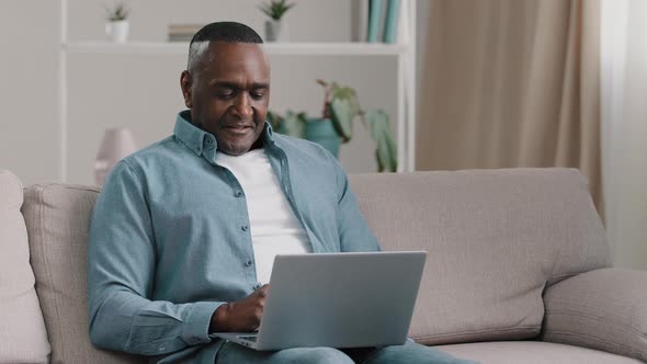 Adult African American Man Sitting on Sofa Typing on Laptop Browsing Email Reading Good News Winning