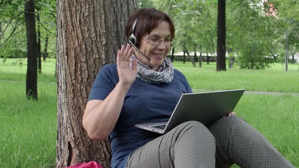 Senior Woman Having On-line Call While Sitting on the Grass in City Park. Education Concept.