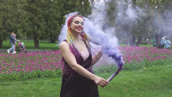 A Girl with a Smile with Bright Makeup and Colored Braids in a Purple Dress Blows Artificial Purple