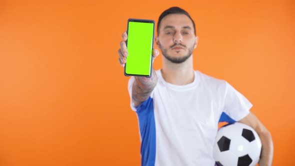 Football Fan In White Blue Shirt and Ball Show Green Screen Smartphone Orange Background 