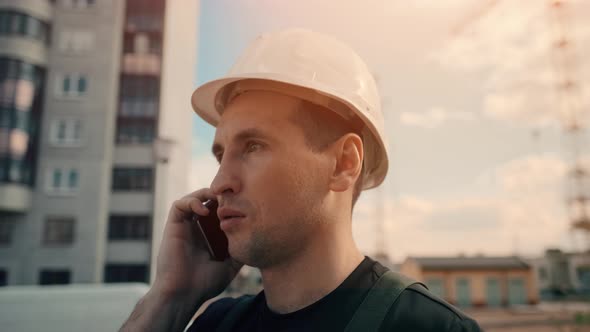 A Builder Wearing Helmet Is Talking By Phone While Inspecting Building Site.