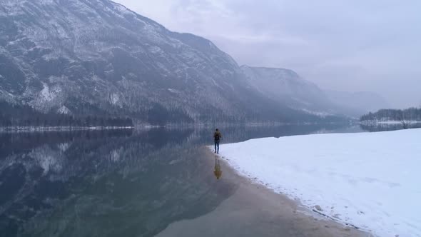 Man walks alone, snowy Bohinj lake reflecting mountains in distance, aerial flyover shot. Slovenia.