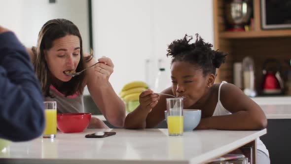 Happy caucasian lesbian couple and their african american daughter eating breakfast in kitchen