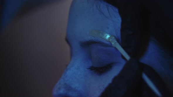 A Man Makeup Artist Waxing the Eyebrow Hairs of His Female Client in Dark Blue Lighting