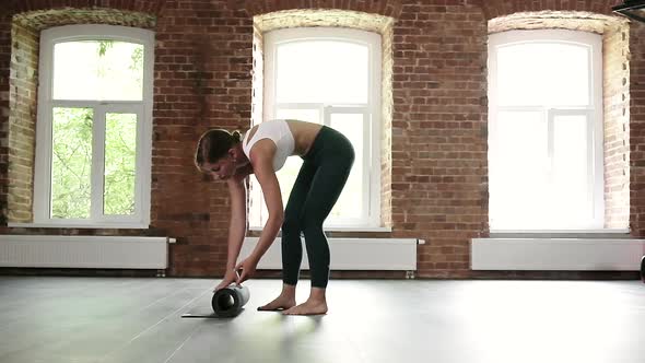 Young Slim Caucasian Woman in Sportswear Rolling Up Yoga Mat on Floor at Gym