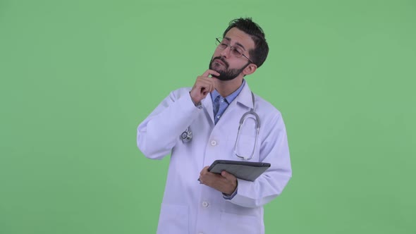 Happy Young Bearded Persian Man Doctor Thinking While Using Digital Tablet