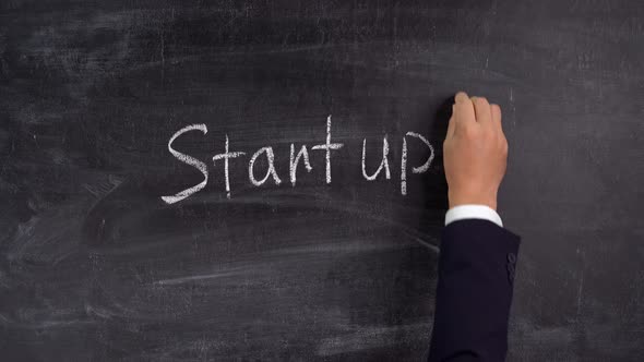 A businessman writes the word STARTUP on a chalkboard and draws an up arrow