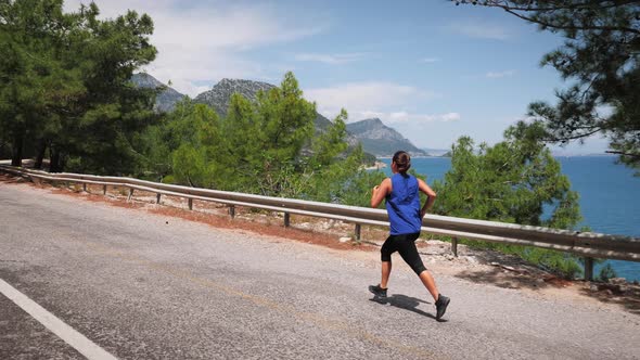 Woman jogging across a highway overpass in mountains with sea on background.