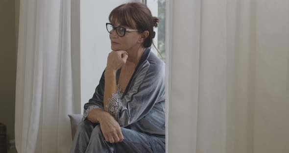 Thoughtful Sad Senior Woman Sitting on Windowsill and Looking Away