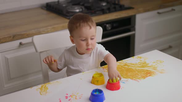 Baby Boy Sitting Behind Table and Drawing with Fingers Covered in Colorufl Paint