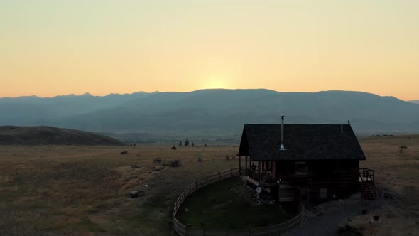 Aerial, American farm house wooden cabin in rural fields during golden hour. Mountain hills in backg