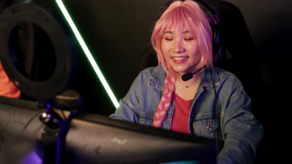 Focused Asian Woman in Her 20s with Pink Hair Participating in an Online Game Sitting in Front of a