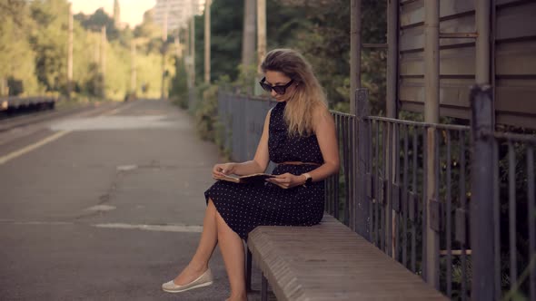 Tourist Reading Book When Waiting Train On Electric Train Station.Girl Relaxing And Sitting On Bench