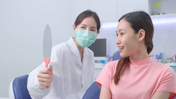 Asian orthodontist holding the mirror to let young girl patient check her orthodontics in clinic.