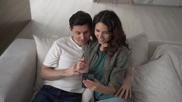 Young Couple in Love Make Online Purchases Using Smartphone While Sitting on the Sofa at Home in the