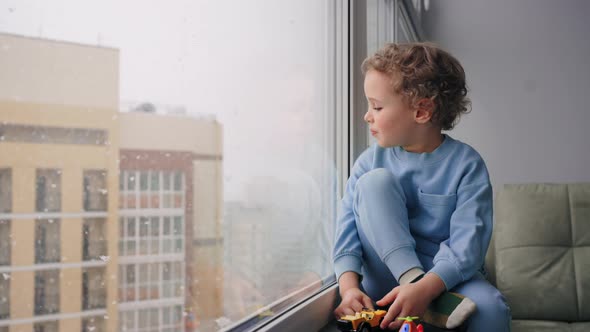 Adoption of Children and Parenting Little Boy is Sitting Alone in Room and Looking on Falling Snow