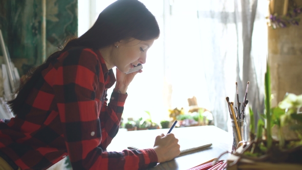 Young Woman Artist Painting Scetch on Paper Notebook with Pencil. Girl Smile and Talks Phone