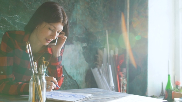 Young Woman Artist Painting Scetch on Paper Notebook with Pencil. Girl Smile and Talks Phone
