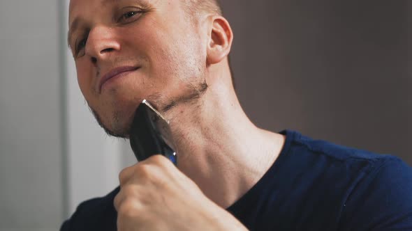 Cheerful Man Shaves Beard with Razor Looking in Large Mirror