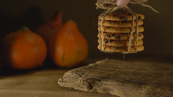 Hand Puts a Pyramid of Chocolate Chip Cookies on a Wooden Table with Pumpkins on a Dark Background