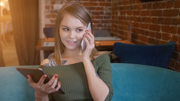 Young Woman Talking on Mobile Phone While Sitting with Digital Tablet on Table in City Cafe,