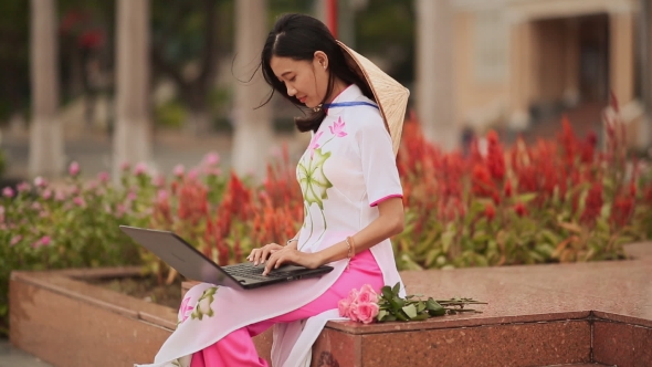 The Beautiful Asian Girl Speaks on a Laptop on the Street and a Beautiful National Dress Ao Dai with