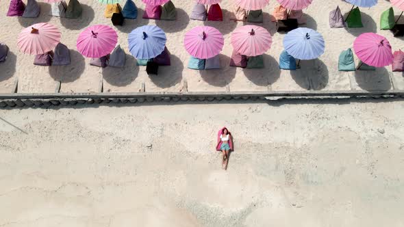 Aerial top down of girl lying and relaxing on tropical beach surrounded by colorful sunshades.