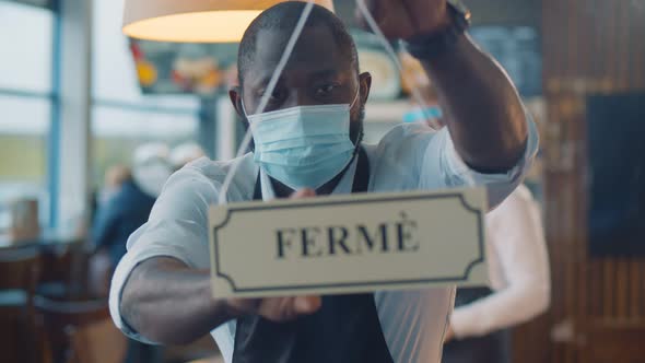 African Waiter in Safety Mask Turning Ouvert Ferme Sign on French Cafe Glass Door