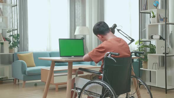 Asian Boy Sitting In A Wheelchair While Using Laptop With Green Screen And Note In Book At Home