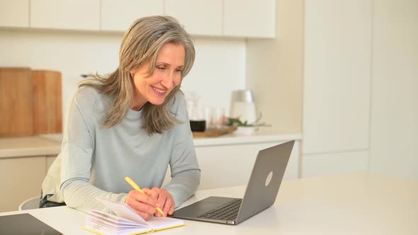 Mature Smiling Woman in Casual Wear Sits at the Desk in Front of Trendy Laptop