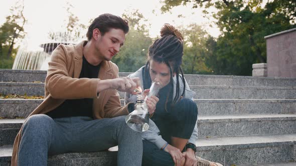 Happy Young Hipster Couple Sitting on Stairs and Smoking Weed with a Bong in Park Slow Motion