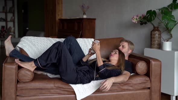 Man and Woman Are Spending Weekend at Home, Lying on Couch in Apartment and Using Smartphone