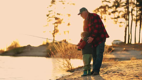 Beautiful Sunset on River Shore Old Fisherman and His Little Son or Grandson are Fishing Together