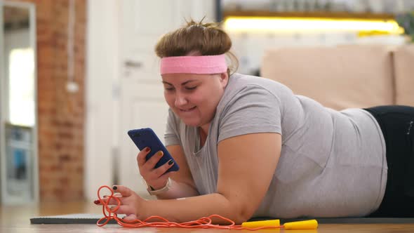 Portrait of Obese Woman Lying on Floor and Using Phone Instead of Training To Lose Weight.