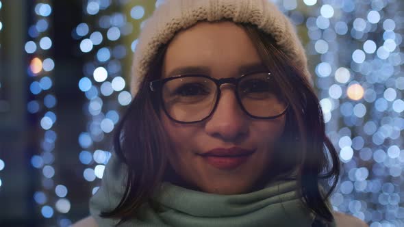 A beautiful smiling brunette in glasses in a winter hat in the night city in the background