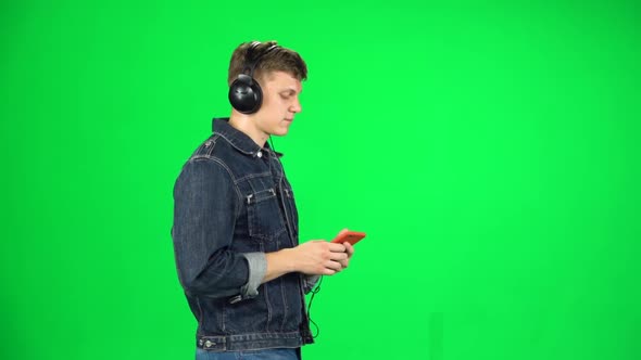 Man in Big Headphones Goes and Texting with Smartphone on Green Screen at Studio. Slow Motion. Side