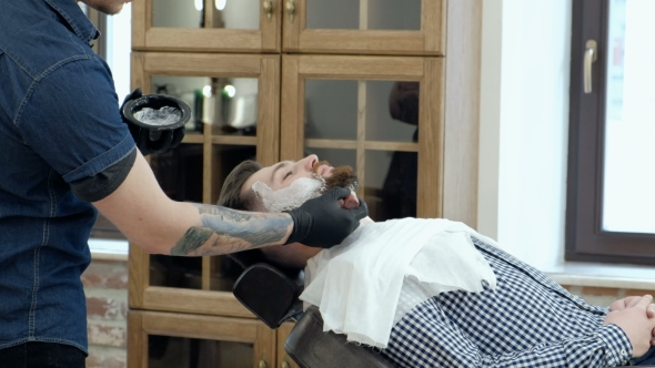 Client During Beard Shaving in Barber Shop. Barber Tattoo on His Arm