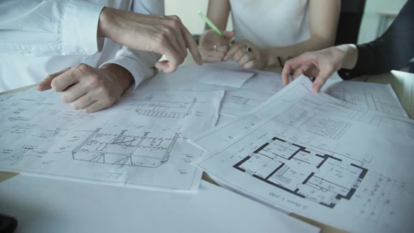 Hands of Three Workers Discussing Building Drawings in Office